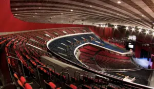 An empty, spacious auditorium with curved rows of red and blue seats, surrounded by red walls and a high ceiling with stage lighting.